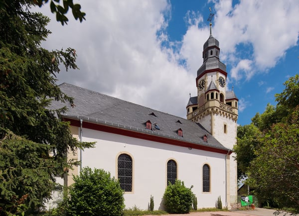Außenansicht des weißen Kirchengebäudes mit schwarzem Dach. Auffällig ist der verzierte Kirchturm.