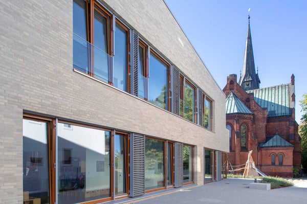 Außenansicht der Kita Hemelingen. Großes, helles Gebäude neben einer Kirche.