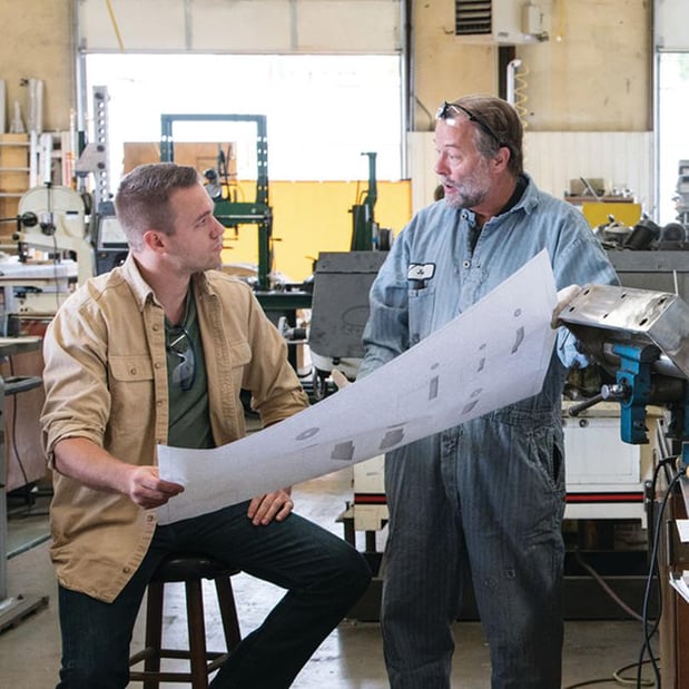 two men in a workshop looking at a blueprint and discussing