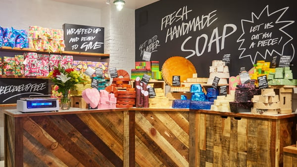 Against a corner of a LUSH store is a large chalkboard sign that says "Fresh Handmade Soap" and in smaller letters "Let us cut you a piece" and on a table sites various colors and sizes of LUSH handmade soaps