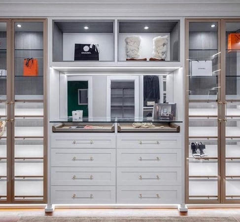 White drawers with mirror above it in closet with gold accessories and lighted cubbies and shoe shelves