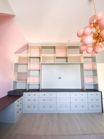 A media center designed in an off white finish with a dark countertop and pink back glass. The closed lower storage contrasts the open upper cabinetry.