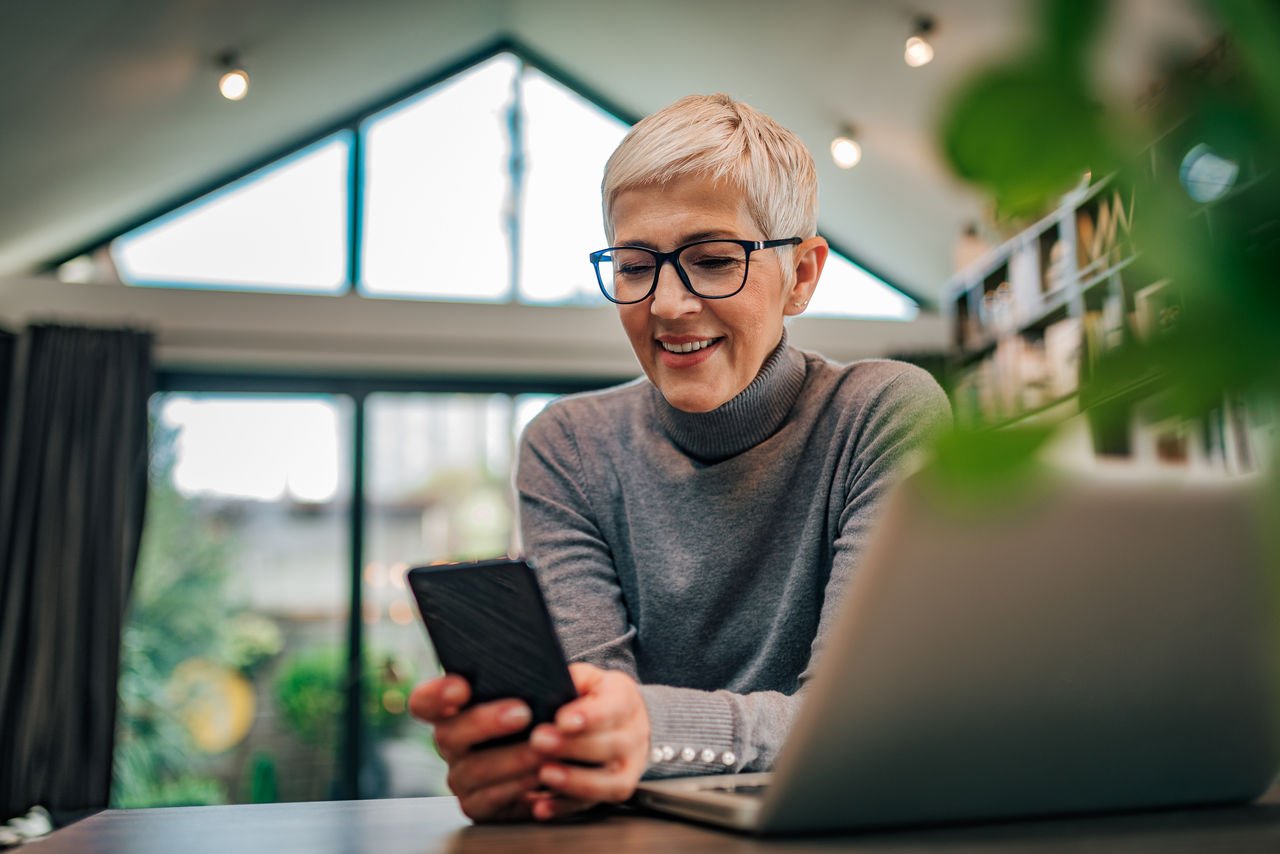 Image of a woman holding her phone smiling, she is sitting in her kitchen with a window behind her and laptop in front of her.