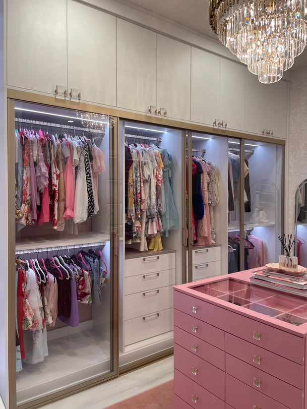 Luxury custom walk-in closet with glass doors, built-in lighting, white cabinetry, and pink island.