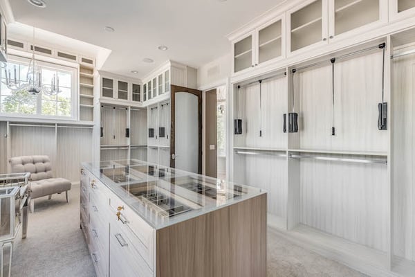 Luxury, custom walk-in closet for her with white wood, large island with glass countertop, and dressing room seating area.