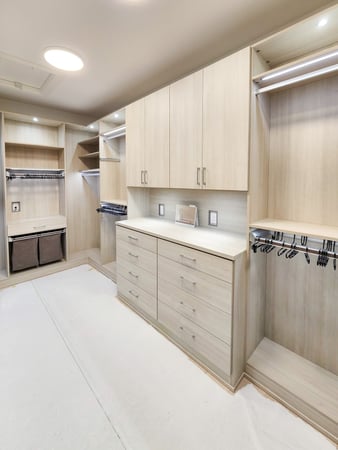 Walk in closet in a light wood grain finish with double hang, upper cabinets, drawers, laundry hampers and lighting.