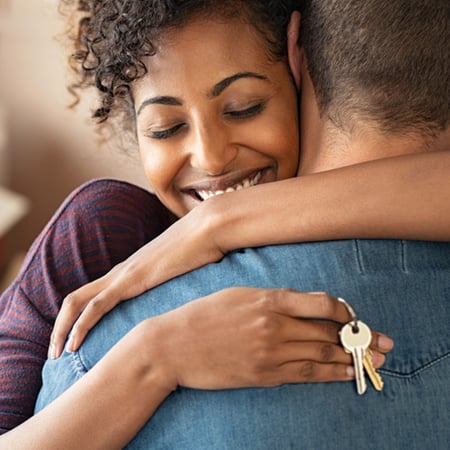 A woman smiles as she hugs her partner while holding the keys to their new home.