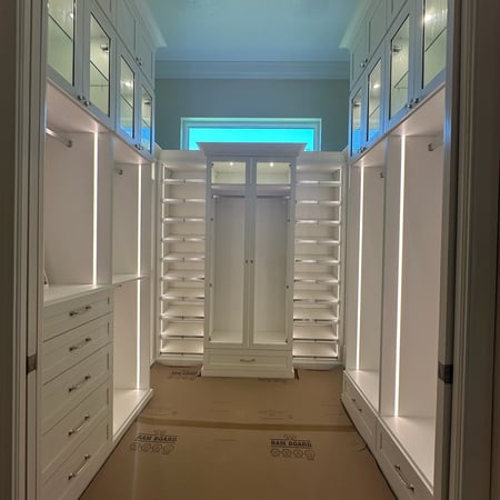Custom master closet in Washed White with polished chrome and nickel hardware, tempered glass shelves, shaker doors with clear glass inserts, microfiber jewelry inserts in Dakota Tan, and integrated LED puck and strip lighting.