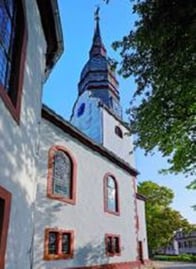 Zu sehen ist die weiße Kirche mit roten Fensterbogen und einem schwarzen Dach. Auffällig ist der spitze Kirchturm.