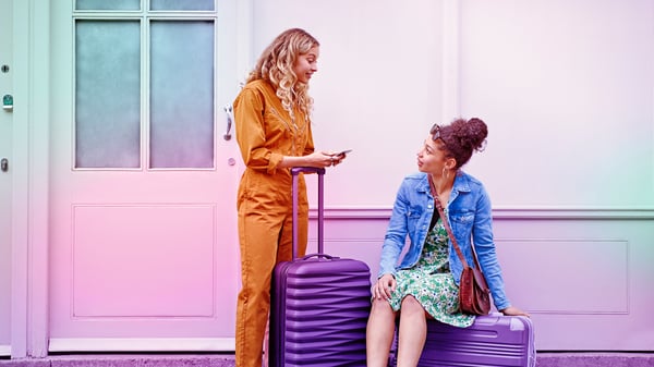 One woman sits on top of a suitcase and another woman leans on the extended handle of another suitcase while she uses her Three Mobile network on her phone.