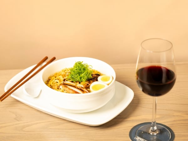 A bowl of Chili Sesame Ramen with chopsticks and a spoon. A glass of red wine on the side.