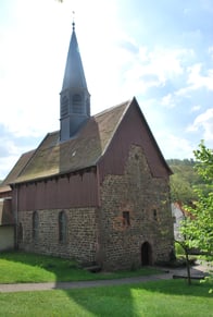Das Bild zeigt die Evangelische Kirche Fraurombach. Sie wurde gebaut aus Backstein und ein spitzer Kirchturm ragt in die Höhe. Die Kirche ist umgeben von einer Wiese und Bäumen.