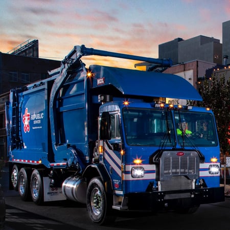A blue dump truck driven by a man in a bright yellow shirt has a red Republic Services Logo on the side
