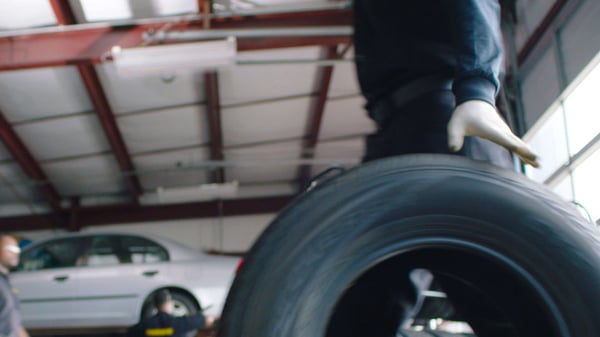 A Meineke mechanic rolls a tire towards a silver car that waits in the background.