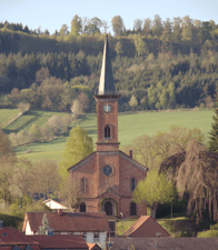 Das Bild zeigt die Außenansicht der Evangelischen Kirche Stockhausen. Die rötliche Kirche fällt durch seinen hohen spitzen Kirchturm auf. Die Kirche ist umgeben von Grünflächen in Form von Wäldern und Wiesen.