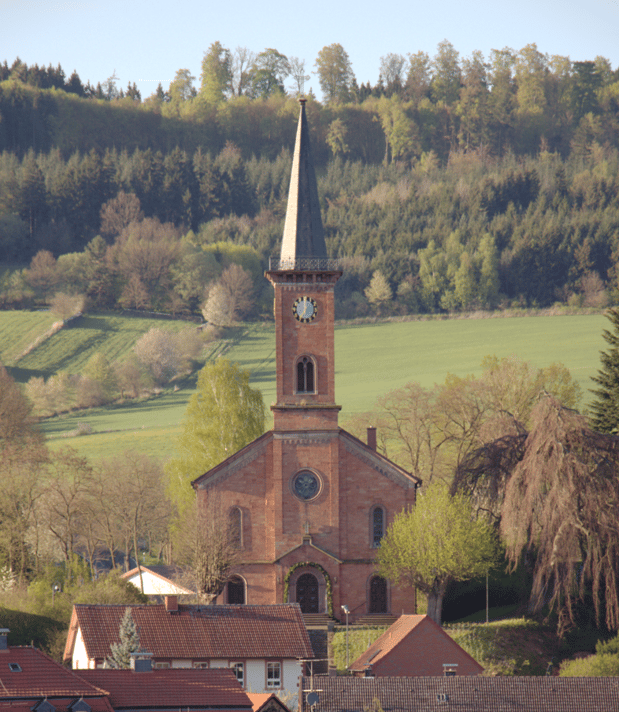 Das Bild zeigt die Außenansicht der Evangelischen Kirche Stockhausen. Die rötliche Kirche fällt durch seinen hohen spitzen Kirchturm auf. Die Kirche ist umgeben von Grünflächen in Form von Wäldern und Wiesen.