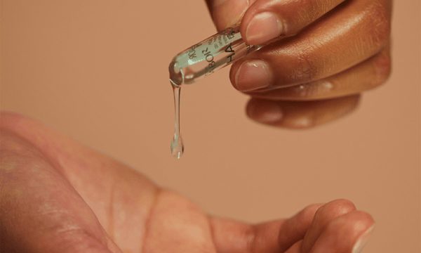 A close-up of a hand pouring a hydrating serum from a glass ampoule into the palm of another hand.
