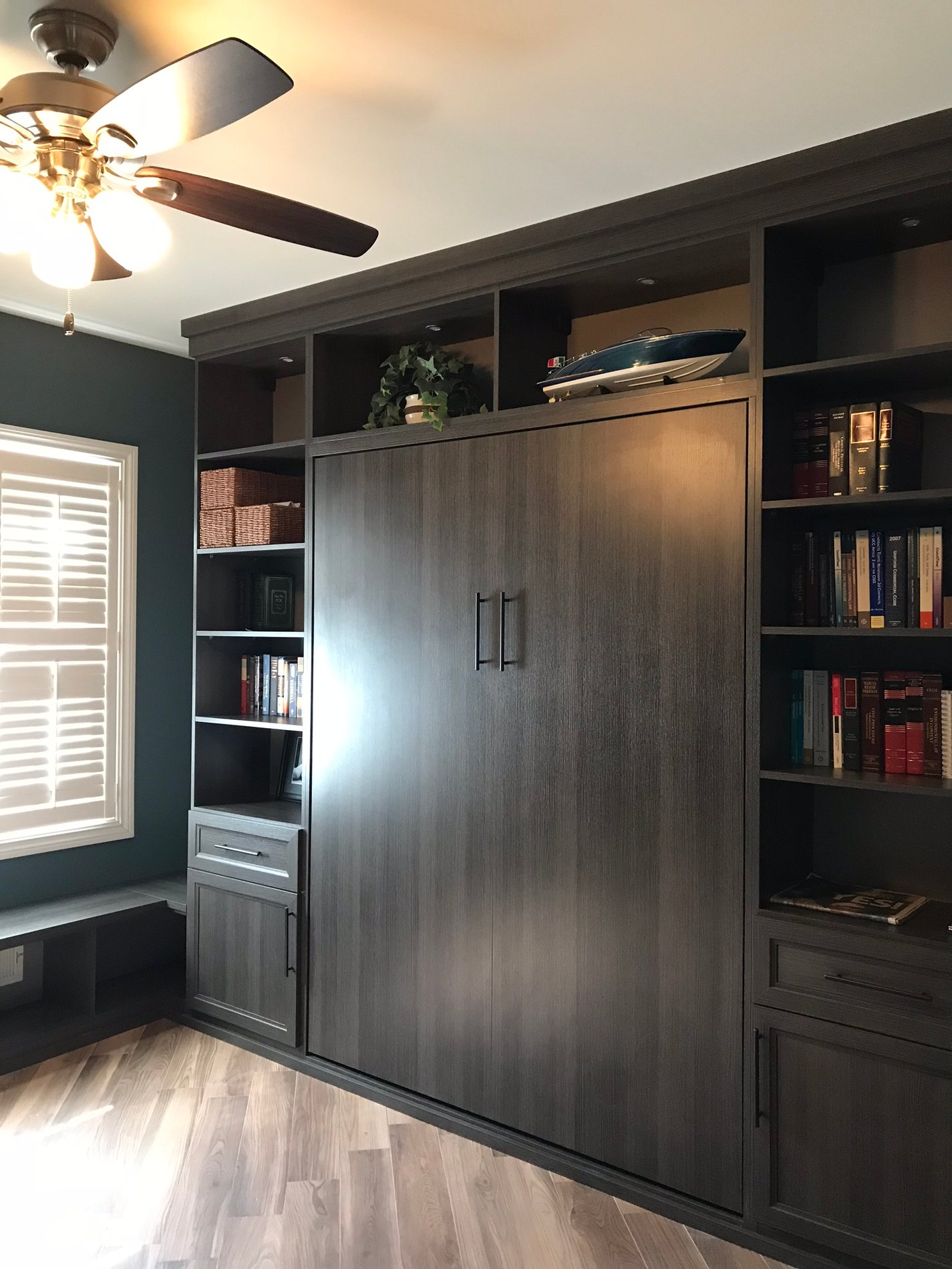 dark grey wall bed folded up with shelving around it, window and ceiling fan