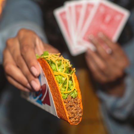 A person holding red playing cards in one hand while offering a Taco Bell Doritos® Loco Taco with the other.
