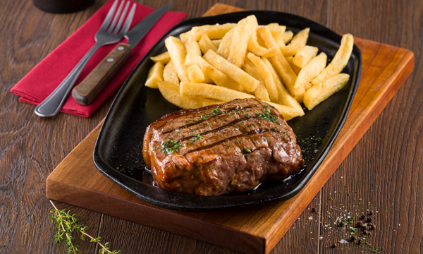 Grilled sirloin steak and chips on a black plate next to a cutlery set.