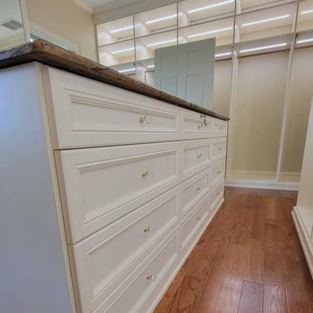Greensboro - Custom closets - Close-up of a custom island with white drawer cabinetry, brass hardware, and hardwood flooring in a walk-in closet.