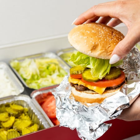 A hand places a bun on top of a cheeseburger in front of a Five Guys catering box featuring all toppings.