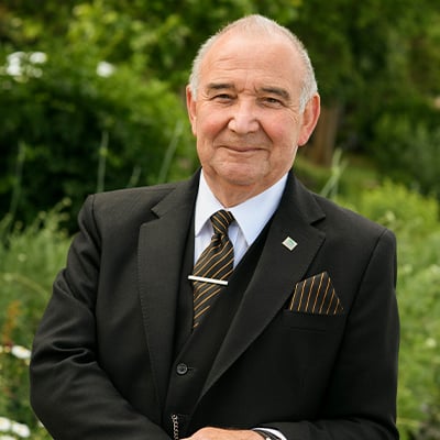 A make funeral director with a black suit and gold and black tie smiles gently to camera