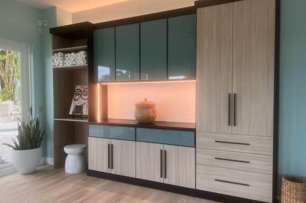 Modern custom built-in with wood-grain and colored cabinetry, illuminated open shelving, drawers, and floor-to-ceiling storage.