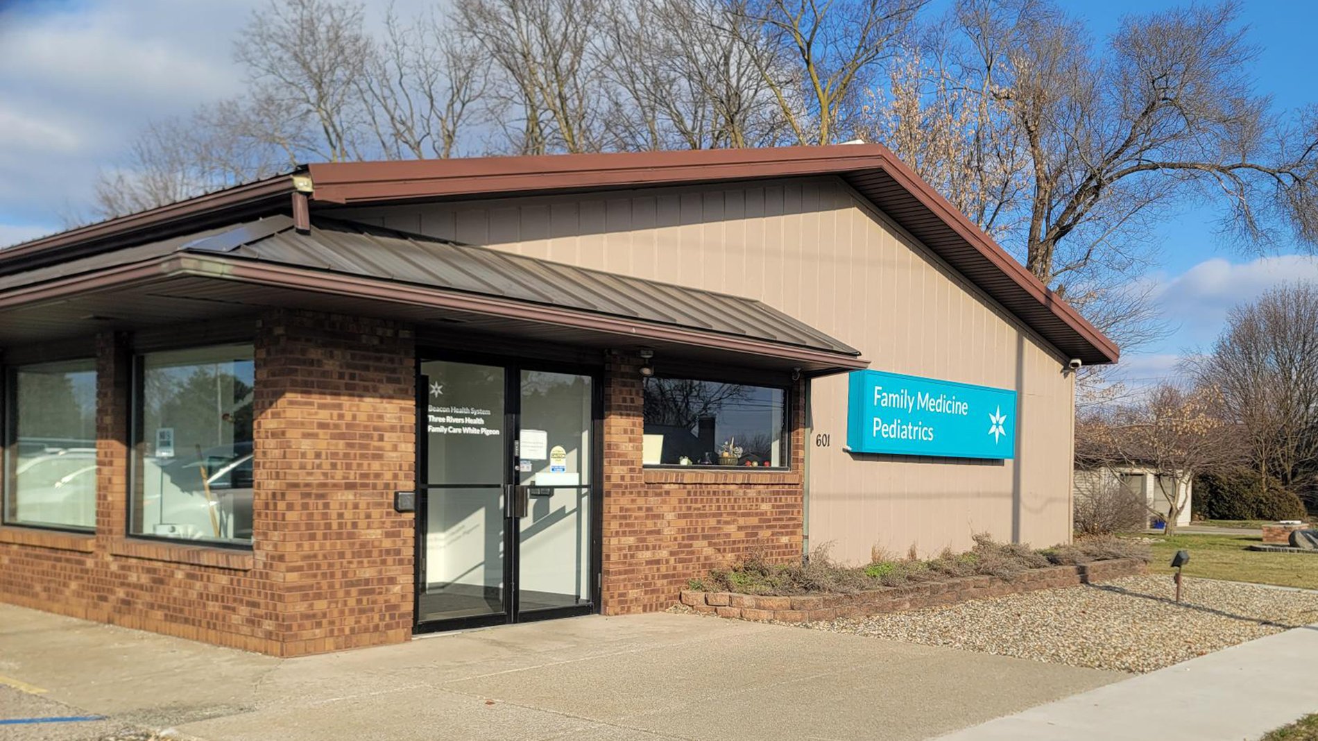 A teal sign stands in the front of the Beacon Three Rivers Health White Pigeon building.  The entrance to Beacon Three Rivers Health White Pigeon with a sign that says "Family Medicine Pediatrics".