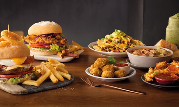 Big Daddy Gourmet Burger with chips, Buttermilk Chicken Gourmet Burger with chips, Asian BBQ Cauliflower Bites, Sesame-Crusted Chicken Strips, and Nachos on a wooden table