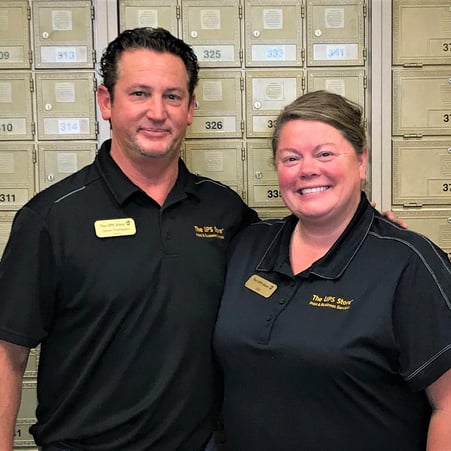 Man and Woman in The UPS Store branded shirts smiling against a wall of mailboxes
