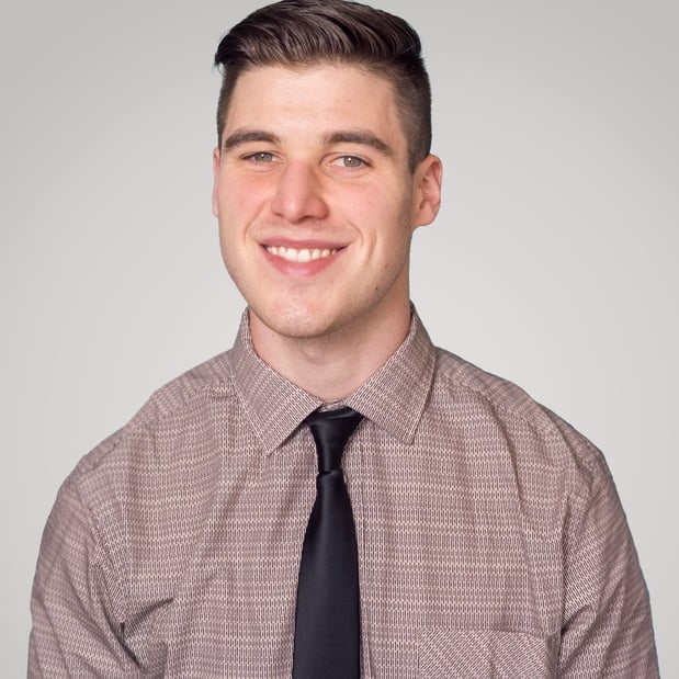Man seated with grey shirt, black tie, and light grey background. 