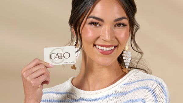 Woman wearing a blue and white striped sweater and fish earrings is smiling and holding up a Cato Fashions gift card.