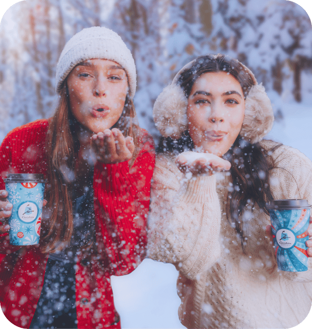 Two women smiling and blowing snow while holding a Caribou Coffee coffee cups.