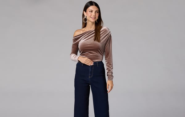 Woman wearing a velvet off the shoulder top and black pants is posing in front of a gray background.