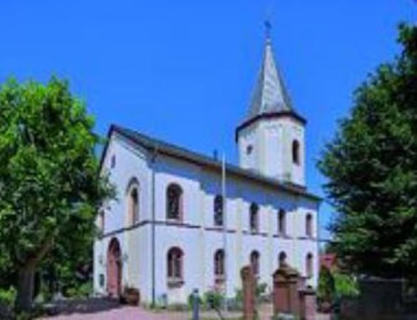 Außenansicht der weißen Kirche. Die Fensterbogen sind rot gehalten. Herausstechend ist der achteckige Kirchturm.