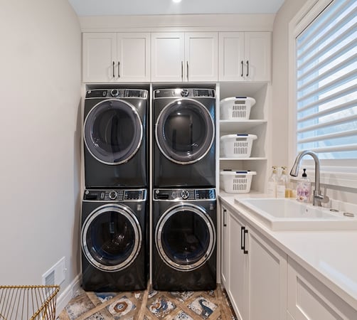 Custom laundry room with stacked washer/dryer, sink and light grey wood cabinets and shelves.