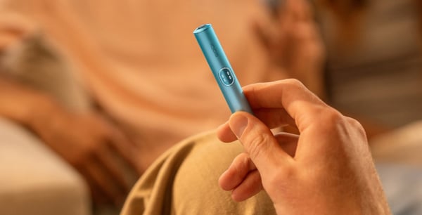 Dłoń trzymająca urządzenie IQOS Iluma na rozmytym tle postaci w beżowej koszuli. Scena oddaje swobodną, ​​swobodną atmosferę.