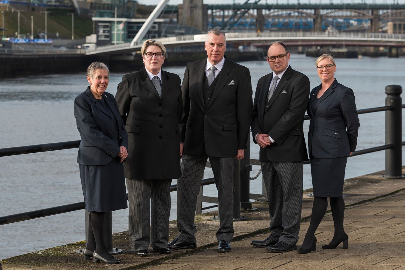 The team from WS Harrison funeral directors in Newcastle stand together on a bridge wearing smart uniforms