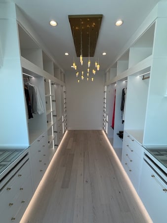 Primary closet in Dove with gold accessories, suspended poles, Lumiere handles, Everstyle jewelry and sunglasses drawers, pullout pant racks, stacked system design, low-iron clear glass countertops, and integrated lighting.