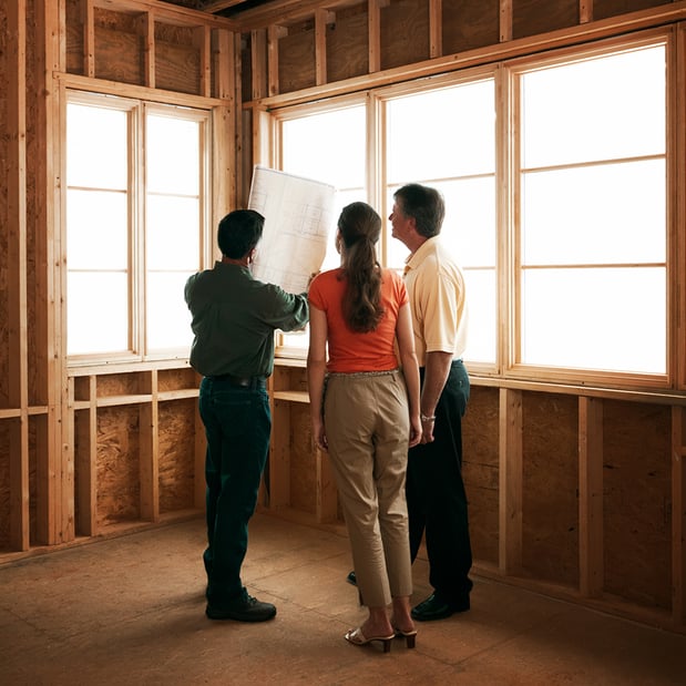 A man holding up building plans to a man and woman in a building that's under construction