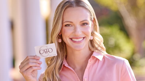 Woman wearing a pink button down shirt and pink drop earrings is smiling and holding a Cato Fashions gift card.