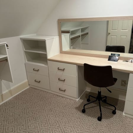 Greensboro - Custom closets - A built-in vanity with drawers, shelving, and a large framed mirror, paired with a desk chair in a softly lit, carpeted room