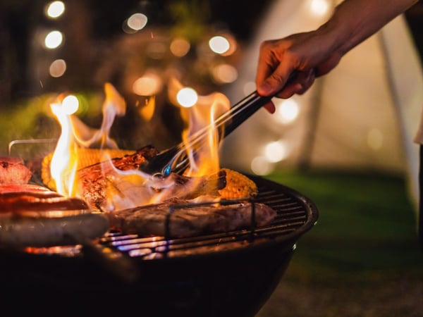Personne cuisinant sur un barbecue au charbon en extérieur avec des flammes, du maïs et des viandes grillées, ambiance estivale conviviale – barbecue disponible chez Boulanger Toulouse Fenouillet.