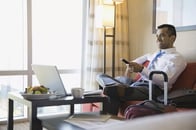 Business man sitting on a couch inside a hotel holding a remote while watching television with his laptop and luggage.