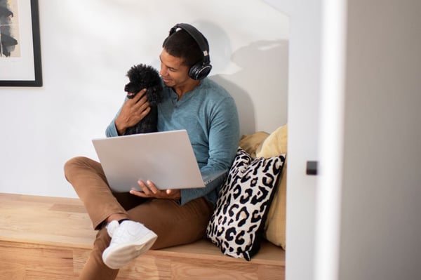 Hombre trabajando dese su hogar con una computadora portátil mientras acaricia a su perro