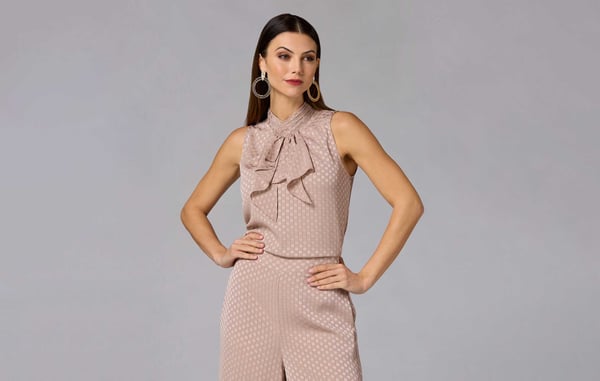 Woman wearing a beige sleeveless blouse and matching beige pants is posing with her hands on her hips in front of a gray wall.