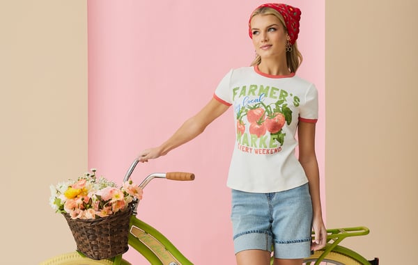 Woman wearing a farmer's market graphic tee and cuffed jean shorts is posing with a green bicycle in front of a pink background.