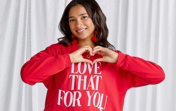 Woman wearing a red sweatshirt is posing with her hands in a heart shape, standing in front of a white curtain.
