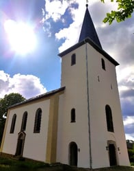 Die weiße Kirche mit spitzem Turm von  der Evangelischen Kirche Alsbach im Westerwald erscheint vor blauem wolkigen Himmel
